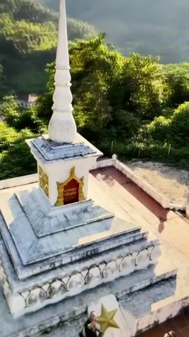 The video shows an aerial view of a white stupa with a pointed spire, set against a backdrop of lush green hills. The stupa is ornate, with tiered levels and decorative elements, including a golden star on its base. A person in dark clothing is visible standing near the base of the stupa. The camera angle shifts, creating a sense of movement and height, and the video ends with a bright, blurred effect.