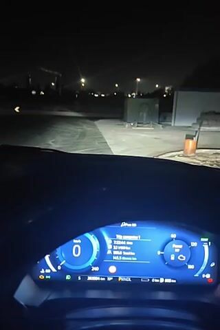The video shows the interior of a car at night, likely parked or driving very slowly. The dashboard is visible with a digital instrument cluster displaying speed, fuel level, and other car information. The infotainment screen shows a radio interface with station options. The outside environment is dark, with street lights and buildings visible in the distance. The car's headlights illuminate the road ahead. The car is not moving or moving very slowly.