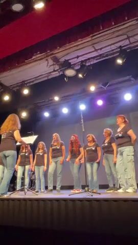 A choir director stands on the left side of a stage, facing a group of eight women singers. The director, wearing a dark t-shirt and jeans, holds a music stand and appears to be directing the singers. The singers, all wearing matching dark t-shirts with a light blue logo and light blue jeans, stand in a line across the stage. They are all looking towards the director and appear to be singing, with some of them holding their mouths open. The stage is lit with bright spotlights, and a red curtain is visible at the top of the frame. A drum set is partially visible behind the singers on the left side of the stage. The overall environment suggests a performance or rehearsal setting.
