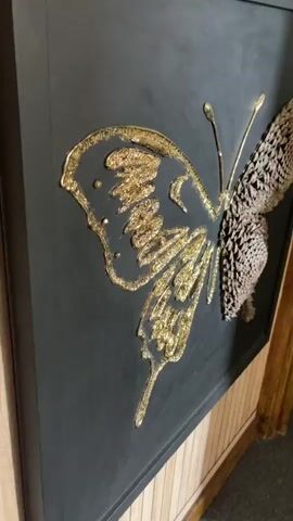 This is a close-up, vertical video of a butterfly artwork. The artwork is mounted on a dark, possibly black, background. One wing of the butterfly is outlined and filled with a shimmering gold material, appearing to be glitter or gold leaf, with a textured, almost three-dimensional effect. The other wing is meticulously constructed from what appear to be small, overlapping natural elements, possibly dried flower buds or pine cone scales, arranged in a circular pattern that mimics the texture and shape of a butterfly's wing. A thin, gold-colored wire forms the antennae and body of the butterfly.

The video pans slightly, showing the artwork from different angles. The background behind the artwork appears to be a wall with vertical wooden panels, suggesting an indoor setting. The lighting is consistent throughout the video, with no obvious time of day indicated. There are no people visible in the media file. The overall impression is of a handcrafted, artistic piece. The location context of Portsmouth, United Kingdom, does not provide any additional visual cues within the video itself. No text is visible in the video.