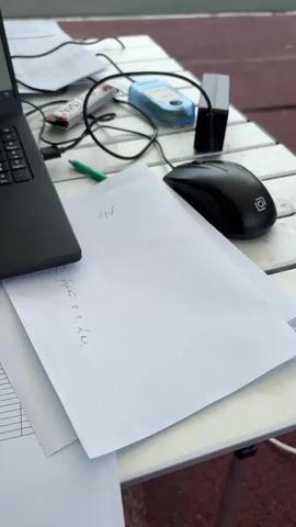 A close-up video shows a table with a laptop, papers, and equipment, likely for a sporting event. The laptop, a black "Aikor" model, displays a spreadsheet. The papers include lists of names and numbers, seemingly related to competitors. There's a black computer mouse, a green pen, and several wristbands with timing chips (indicated by red tags and numbers) on the table. The environment appears to be outdoors, with a glimpse of a maroon-colored surface and a tent-like structure in the background. The table is white and has a folding leg, suggesting a portable setup.