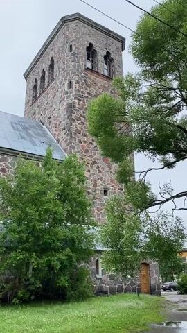 The video shows a stone church with a tall bell tower. The church is made of large, rough-hewn stones, and the bell tower has arched windows at the top. There are trees and grass in the foreground, and the sky is overcast. The camera pans slightly to the left, revealing more of the church's stone structure and a portion of its metal roof. The scene is static, with no visible movement apart from the camera's slight pan.