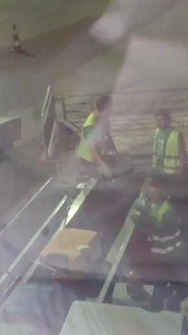 The media file depicts a scene at what appears to be an airport or similar transportation hub, likely at night given the artificial lighting. In the foreground, a conveyor belt system is visible, carrying luggage. Two individuals wearing high-visibility vests are interacting with the luggage. One individual, wearing a bright yellow vest, is standing and appears to be attending to a piece of luggage on the belt. Another individual, in a green vest, is further down the belt and also seems to be working with the luggage. In the background, another person in a green vest is visible. A safety cone is present in the upper left corner of the frame, suggesting an operational area. The overall atmosphere is functional and appears to be related to baggage handling. The quality of the footage is grainy and somewhat blurred, possibly due to low light conditions or the recording method. No visible text is present in the media file. The setting in Mouzouras, Greece, would imply an outdoor environment, possibly an apron area of an airport.