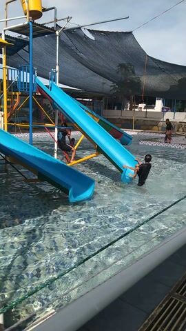In a bright, outdoor setting, likely a water park, several children are enjoying a day of fun. The primary focus is on a blue water slide structure in a swimming pool. One child, wearing a black outfit, is positioned at the top of the slide, about to go down. Another child, also in black, is visible further down the slide, splashing into the water. A third child is in the pool near the slide's exit. The water is clear and reflects the sunlight, indicating a sunny day.

The water slide itself is a vibrant blue, supported by a colorful metal frame of yellow, red, and blue. Above and behind the slide, a large black mesh net is suspended, likely to provide shade. In the background, there are buildings with white walls and blue accents, suggesting changing rooms or other park facilities. A few other individuals can be seen in the distance near the edge of the pool and on a walkway.

The overall mood is one of cheerful recreation and summer enjoyment. The time of day appears to be late morning or early afternoon, given the bright lighting and clear sky. The location cues, such as the architecture and the lush greenery in the background (though partially obscured by the netting), hint at a tropical or subtropical climate. There is no visible text in the media file.