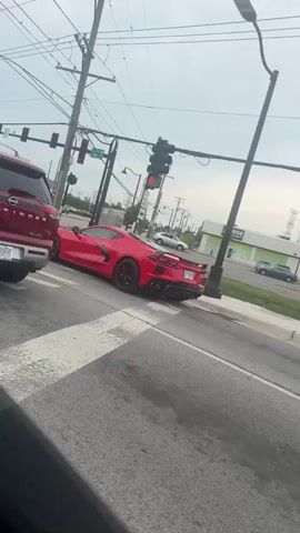 A red Chevrolet Corvette is driving down a street. The car is a mid-engine sports car with a sleek, modern design. It has a large rear spoiler and quad exhaust pipes. The car is driving on a multi-lane road with traffic lights and power lines. In the background, there are businesses and other vehicles. The sky is overcast.