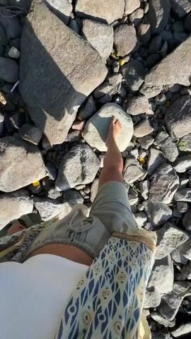 The video shows a first-person perspective of someone walking barefoot on a rocky shore. The subject is wearing light-colored shorts and a white tank top, with a patterned kimono or cover-up draped over their shoulders. The ground is covered with a variety of grey and brown rocks of different sizes, with some sparse green vegetation visible. The sunlight suggests it is daytime, likely late afternoon given the shadows cast by the rocks. The setting appears to be a natural environment, possibly a lake or riverbank, given the rocky terrain. The mood is relaxed and casual, with the focus on the sensory experience of walking on the uneven surface. There is no visible text in the video. While Valaam, Russia is provided as location context, the visual elements alone do not offer specific cues to confirm this location.