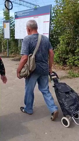 The video begins showing two people walking away from the camera on a paved path next to train tracks. A woman in a floral jacket and dark pants walks ahead of a man in a t-shirt and jeans carrying a bag. The scene then shifts to a sidewalk with a metal fence. An elderly woman with a cane and a backpack walks away from the camera, carrying a bag. The camera pans to show a street with several cars and vans, and a building with signs that read "НАПИТКИ" and "Мариет". The video continues to show people crossing a street at a crosswalk, with pedestrian crossing signs visible. More vehicles are seen in the background, including buses. The video ends with a view of the street and sidewalk.