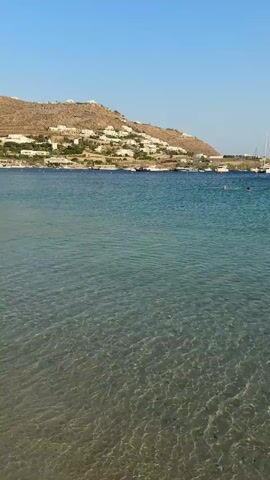 The video captures a serene beach scene in Agios Ioannis Diakoftis, Greece. The foreground features shallow, crystal-clear turquoise water gently lapping against a sandy shore. The seabed is visible through the translucent water, revealing a rippled sand pattern. In the mid-ground, the water deepens to a richer blue, dotted with several boats anchored offshore. A white sailboat with its mast prominently displayed is visible on the right side of the frame.

The background is dominated by a sun-drenched hillside covered with white buildings, typical of Greek architecture, cascading down towards the coast. The sky is a clear, vibrant blue, indicating a bright, sunny day. The overall atmosphere is peaceful and idyllic, suggesting a perfect day for relaxation by the sea. The time of day appears to be late morning or early afternoon, given the strong sunlight and clear skies. There are no visible people in the immediate foreground, but a few specks in the water and on the distant shore might indicate swimmers or beachgoers. No text is visible in the media.