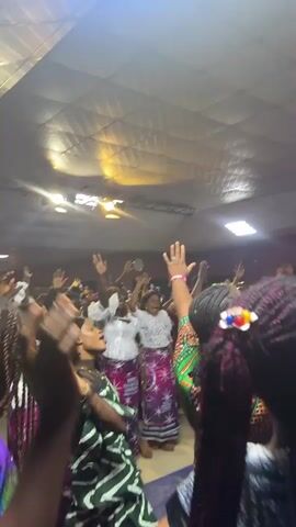 The video shows a group of people raising their hands in what appears to be a religious or celebratory gathering. The lighting is dim, with bright spotlights on the ceiling. The individuals are dressed in colorful clothing, some wearing African prints. Their faces are turned upwards, and their expressions suggest emotion and devotion. The camera is shaky, indicating movement and a dynamic atmosphere. The overall scene conveys a sense of community and shared experience.