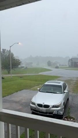 The video shows a view from a porch looking out at a street during a rainstorm. A silver car is parked in the driveway. The street is wet and rain is falling. There are houses and trees visible in the background, shrouded in a misty haze. The sky is overcast and grey.