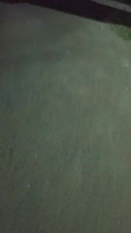 The video shows a dark, outdoor scene at night. A paved surface, possibly a road or sidewalk, occupies the foreground. In the background, there are indications of buildings and streetlights, suggesting a residential or urban environment. The camera appears to be moving, possibly handheld, creating a slightly shaky and dynamic perspective. Shadows are cast across the ground, indicating the presence of light sources, likely from the buildings or streetlights. The overall ambiance is dark and somewhat indistinct due to the low light conditions.