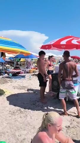 A sunny day at the beach. Several people are visible on the sand and in the water. An ice cream cart with a red and white umbrella is on the beach. Two young men are standing near the ice cream cart. One of them is talking to a woman. In the foreground, a woman is taking a photo with her phone. The water is a bright turquoise color. There are many beach umbrellas and people enjoying the water.