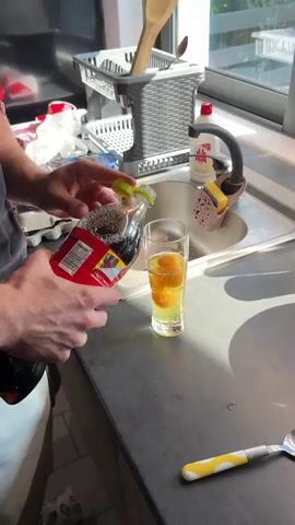 A person's hands are shown pouring cola from a red bottle into a tall glass containing what appears to be raw eggs. The glass is placed on a dark countertop, next to a kitchen sink. In the background, there is a dish rack, a bottle of dish soap, and a sponge. The action takes place in a brightly lit kitchen.