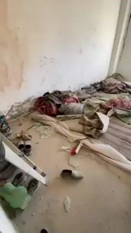 The camera pans across a room showing the aftermath of some sort of damage. The walls are white, but cracked and partially destroyed. There are two windows in the back wall. The floor is covered in debris including cloth, a stripped rug, pieces of plaster, painting tools and materials, and a pair of shoes.
