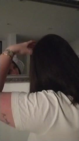 A person wearing a white t-shirt is using a rotating hair brush on dark, straight hair in front of a mirror. The person is facing the mirror and the camera angle focuses on their back and hair. The mirror reflects a portion of the room behind them.