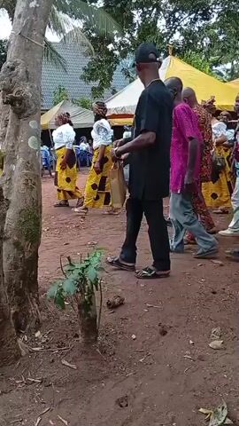 The video shows a group of people gathered outdoors. Most of them are walking around on an area of reddish-brown dirt. Some of them are wearing traditional African clothing. Several tree trunks are visible. There are tents in the background. A man with a black baseball cap is walking in front. The sound of African music is heard.