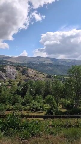 The video shows a panoramic view of a mountainous landscape on a sunny day with scattered clouds. In the foreground, there is a fence and vegetation. The midground features a densely wooded area with various trees. Beyond the trees, the terrain rises into hillsides dotted with buildings and rocky outcrops. The background is dominated by rolling mountains under a bright blue sky with fluffy white clouds. The camera pans slightly, revealing the vastness of the natural environment. No people or animals are visible. The scene appears tranquil and picturesque.