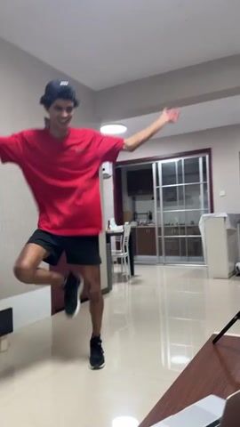 A young man dances in a living room, wearing a red t-shirt, black shorts, black athletic shoes, and a black baseball cap backwards. He faces toward the camera in the first frame and turns toward his left in the third frame. He moves across the floor, apparently practicing dance moves. A laptop is open on a wooden coffee table in the foreground. A camera on a tripod stands next to the table, indicating he is filming himself. The room is sparsely furnished and has tiled floors. A kitchen is visible through a doorway and beyond a sliding glass door. Other doorways lead to other rooms in the apartment. A modern light fixture is visible in the ceiling.