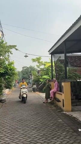 A man rides a cream-colored scooter down a narrow, paved alleyway. He is wearing a yellow t-shirt and black shorts. To his right, two women are seated on a low yellow wall. One woman wears a green dress and the other wears a pink patterned outfit and a headscarf. There are trees and houses visible in the background. The sky is overcast.