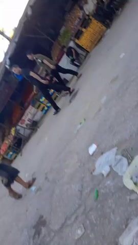 The video shows a street market scene. In the foreground, a person's legs are visible, wearing black pants and white shoes. They are holding a white bag with a red design. In the background, several people are walking and standing near market stalls. The stalls appear to be selling fruits and other goods. The environment looks like a typical outdoor market with awnings and structures overhead. The lighting suggests it is daytime. The camera perspective is low and tilted, giving a dynamic and slightly disorienting view of the scene. There is debris on the ground in some areas.
