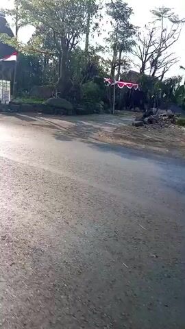 The video shows a paved road with a motorcycle passing by. On the left side of the road, there's a traditional-looking building with a conical roof, possibly a hut or a ceremonial structure. There are also some flags and a sign. A person is seen riding a motorcycle from left to right, followed by another motorcycle carrying two people. The environment appears to be rural with trees and vegetation visible. The sky is bright, suggesting daylight.