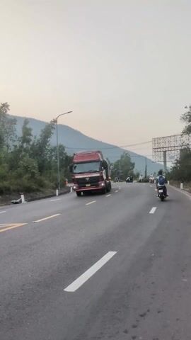 The video shows a two-lane asphalt road winding through a mountainous landscape at what appears to be dusk or dawn. A large red semi-truck with a long trailer, covered by a dark tarp, drives past the camera from left to right. The text "ĐÔNG NAM Á" is visible on the cab of the red truck. Following the red truck, a white truck with a covered bed drives in the same direction. Several motorcycles with riders are also visible on the road, both ahead of the trucks and driving past the camera.

The road features white lane markings. On the right side of the road, there's a road sign with "10%" written on it, likely indicating a gradient. Further down the road on the right, there are large billboard-like structures. Trees and foliage line both sides of the road, and mountains are visible in the background against a light, overcast sky. A streetlight is also visible on the left side of the road. The scene depicts typical traffic in a semi-rural, hilly area.