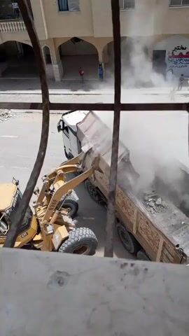 From behind a window grate, the video shows a CAT front loader dumping a load of demolition debris into the back of a dump truck. The loader is on the left of the scene and the truck is on the right. The dust from the demolition debris obscures the view. The truck is parked on a paved street with parking lines. The location appears to be an urban area in the Middle East, as a building with arched doorways can be seen in the background. Two people are standing beneath one of the arches. One of the people is a child.