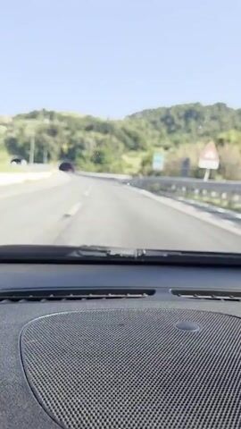 The video is filmed from the dashboard of a car driving on a highway. The road ahead is paved and has lane markings. There are guardrails on the right side of the road and a concrete barrier on the left. In the distance, there is a tunnel entrance. The sky is clear and blue, and the surrounding landscape is hilly and covered with green trees. The sun is shining, creating a bright and clear scene. The video captures the movement of the car as it approaches the tunnel.