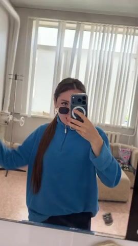 A young woman with long brown hair tied in a ponytail is taking a mirror selfie in what appears to be a bedroom. She is wearing a blue zip-up sweatshirt and black pants. She has sunglasses on and is holding a smartphone in her right hand, taking the photo. Her left arm is raised in the air. The background shows a window with vertical blinds, a couch, and other furniture, suggesting an indoor domestic setting. The lighting indicates it is daytime. The location context provided is "Old Tuymazy, Russia."