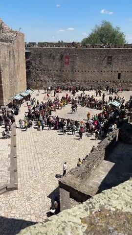 The video shows a crowd of people gathered in the courtyard of a castle on a sunny day. In the center of the courtyard, two people dressed in medieval armor are engaged in a staged sword fight, surrounded by a wooden railing. Numerous vendors have set up stalls around the perimeter, and people are milling about, watching the fight and browsing the goods. The castle walls, made of large stone blocks, rise high above the courtyard, with banners hanging from the walls. The ground is paved with cobblestones.