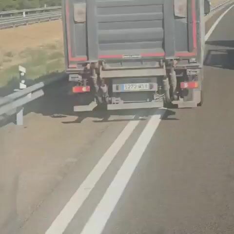 The video shows a close-up view of a concrete barrier on the side of a road, likely a highway. The barrier is gray and appears to be made of several modular sections. The surface of the concrete is slightly weathered, with some dirt and debris. In the background, a road with white lane markings is visible, separated from the barrier by a patch of dry, grassy terrain. Beyond the road, there's a slight incline with more vegetation. The overall lighting suggests daytime, possibly bright sunlight, casting shadows.