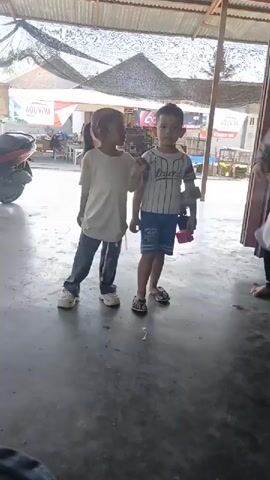 Two young children, a boy and a girl, stand facing the camera in what appears to be an outdoor covered area. The girl, on the left, wears a white t-shirt and gray pants. The boy, on the right, wears a striped t-shirt and blue shorts, and is holding a toy gun. The floor is concrete. In the background, a motorcycle is visible to the left, and a red awning is above.