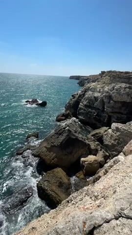 The video pans across a rocky coastline on a sunny day. The turquoise ocean water crashes against the shore, creating white foam. Jagged rocks and cliffs form the edge of the land. In the distance, the coastline stretches out, and a few people can be seen standing on the cliff edge. The sky is a clear blue.