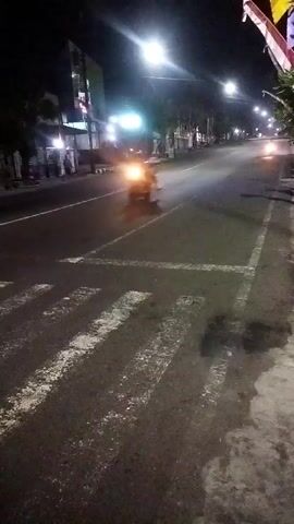 The video shows a nighttime street scene. A single motorcycle is visible in the middle of the road, moving away from the camera. The road has a crosswalk with white stripes painted on it in the foreground. Streetlights illuminate the scene, casting long shadows. Buildings line the sides of the road, with some lights on. The overall impression is of a quiet, sparsely populated urban or suburban street at night.