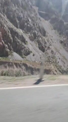 The video shows a view from a moving vehicle, likely a car, driving along a road. A large, rocky mountain rises alongside the road, with patches of sparse vegetation. Power lines and a large electrical tower are visible against the backdrop of a blue sky with scattered clouds. The road is paved with a solid white line marking the edge. The sunlight is bright.
