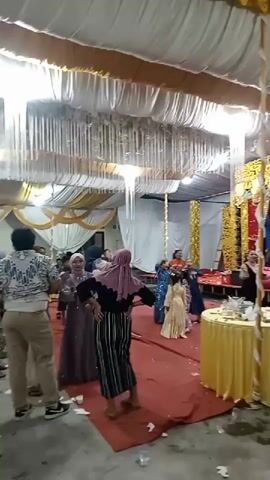 A wedding reception is in progress, with guests dressed in formal attire, some wearing hijabs. A stage is set up with a banner that reads "ANISSA SUHAIMI." A female performer is on stage, singing and dancing. There are several chafing dishes filled with food, suggesting a buffet. The room is decorated with white and gold drapes and fabric, and there are strings of lights hanging from the ceiling. Some confetti is scattered on the red carpeted floor.
