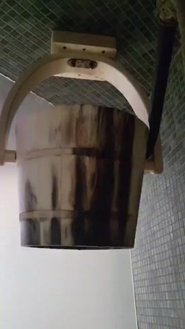 A brown and white bucket is shown suspended by a white wooden frame and support structure. The bucket tips over and dumps out water. The water streams down and then cuts off.  The scene is in what appears to be a spa, as the wall is tiled with square tiles and the ceiling has a green wire mesh covering.