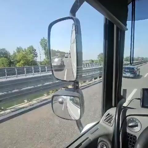 The video shows a view from inside a truck cab, looking forward and into the side mirrors. The truck is driving on a highway. In the side mirror, another car is visible. In the main view, the road ahead is visible, with other vehicles including cars and trucks. The weather appears to be clear and sunny. The truck has a GPS device mounted on the dashboard.