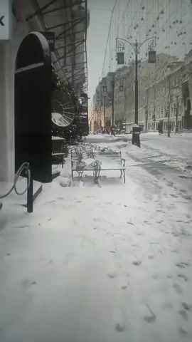 A wintry street scene is captured in a slightly grainy video. The sidewalk is heavily covered in snow, with footprints scattered across its surface. A metal bench stands to the right, also draped with snow, and adorned with decorative metal figures in human-like poses.

To the left, the front of a building is visible, decorated for the holidays with greenery and ornaments. A sign reading "BRO&N" is visible.  A large, round, metallic decoration hangs near the front of the building.

In the distance, the street is lined with buildings, and street lamps are suspended overhead. Several people can be seen walking in the distance, and it appears to be a cold, snowy day.