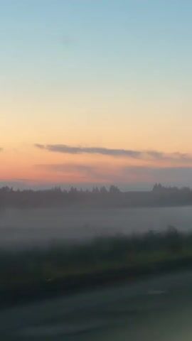 The video shows a landscape at dawn or dusk. The sky is a gradient of blue and orange, with faint clouds. In the middle ground, there is a body of water or a low-lying area with mist or fog. A dark line of trees forms the horizon. In the foreground, a road is visible, with cars driving on it. One car's headlights are on. The camera appears to be moving, possibly from inside a vehicle.