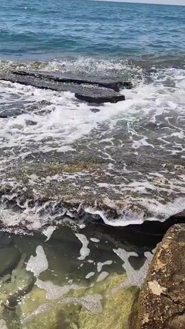 The video shows a close-up view of ocean waves crashing against dark, flat rocks on a sunny day. The water is clear and slightly greenish, with white foam from the waves. The sky is blue with a few scattered clouds. The camera pans slightly to the right, revealing more of the rocky shoreline and the expanse of the ocean.