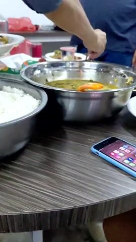 A person serves themselves food from a buffet-style arrangement. There is a large bowl of white rice, a bowl of soup with what appears to be a large carrot slice, and a plate of sauteed vegetables. The food is on a dark wood-patterned table. A smartphone with a blue case sits on the table next to the food. Other people are visible in the background, and additional plates of food are also visible.