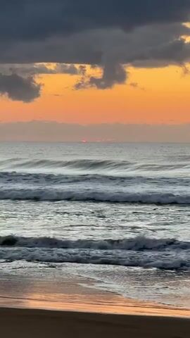 The video shows a serene beach scene at sunrise. The sky is a gradient of dark grey clouds above, transitioning to a vibrant orange and pink hue near the horizon. A sliver of the sun is visible just above the ocean's surface, creating a faint glow. The ocean is calm, with gentle waves rolling towards the shore, their foam glistening. The wet sand in the foreground reflects the colors of the sky and the subtle glow of the rising sun. No people or objects are present in the frame, creating a peaceful and undisturbed natural landscape.