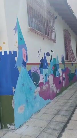The video shows a mural painted on the exterior wall of a building. The mural features cartoonish castles and a figure wearing a wizard hat, all in bright colors. To the left of the mural, a flagpole with a Greek flag is visible. The wall appears to be slightly damaged in some areas, with peeling paint. The ground in front of the wall is paved with light-colored tiles, and some greenery is present on the left side of the frame. The building has a tiled roof and barred windows. The lighting suggests it is daytime.