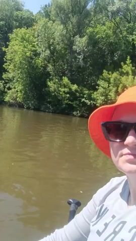 The video begins with a close-up of a person wearing an orange hat and sunglasses, looking towards the camera. The person is in a body of water, possibly a river or lake, with lush green trees lining the bank in the background. The water appears murky and brown. The perspective then shifts to a wider shot, showing the person from the waist down, lying on their back in what appears to be an inflatable raft or kayak. Their legs are extended, and they are wearing blue shorts and a grey long-sleeved shirt. In the distance, another person can be seen on a paddleboard, also in the water. The video continues to show a similar scene, with the camera person's legs visible and the paddleboarder in the background, before cutting back to the initial close-up of the person in the orange hat. The overall setting is a calm, sunny day outdoors, with emphasis on water activities and nature.