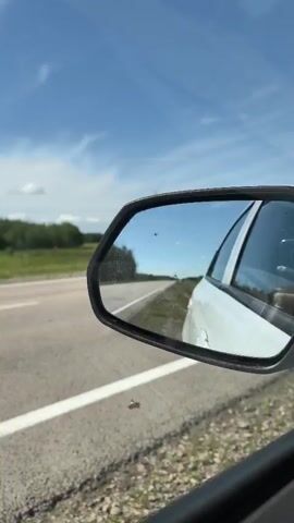 A rearview mirror reflects a sunny day with a blue sky and scattered clouds. The mirror shows a white car, a road with a white line, and trees in the distance. The car is moving, as indicated by the slight blur of the landscape in the mirror.