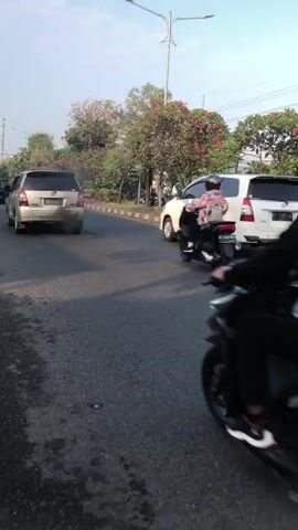 A white SUV is parked on the side of a road, facing forward. A motorcycle is approaching the SUV from behind and to the left, with a rider and passenger on board. The rider is wearing a helmet and the passenger is wearing a brightly colored shirt. There are trees and other vegetation on the right side of the road. The sky is clear and blue.