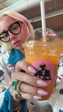 The video shows a person with blonde hair and glasses holding a clear plastic cup with an orange drink and a straw. The person is wearing a white jacket with blue accents and several rings on their fingers. The cup has a pink circular sticker with a silhouette of a man holding a cup. The person brings the straw to their mouth and takes a sip of the drink. The background appears to be indoors, with some industrial-looking elements visible, such as a pipe and a metal grate. The lighting is bright, suggesting daylight or well-lit indoor conditions.