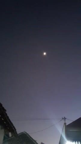 This is a vertical, handheld video of a moonlit night sky over a residential area. The moon is visible as a bright, small orb in the upper middle portion of the frame. Below the moon, the sky is a deep, muted purple, transitioning to a lighter lavender at the bottom. The silhouettes of several rooftops and buildings are visible in the lower half of the frame. The roof on the left has a tiled surface. In the center and right, there are white buildings with visible windows and a porch area. A bright light source, possibly from a streetlamp or a porch light, is illuminating the right side of the frame, casting a lens flare. There are power lines visible horizontally across the frame. The overall impression is a quiet, nighttime scene in a suburban or urban environment.