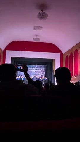 A video shows a school band concert on a stage inside a theater. The camera is positioned in the audience, behind two silhouettes of people, one of whom is holding a camera. The camera zooms in to focus on the stage where a conductor in a suit stands with his back to the audience, directing a group of young musicians wearing matching black uniforms with yellow accents. Behind the band is a banner printed in a foreign language with musical notes in the background. The theater is decorated with a red and white color scheme. The ceiling is lit with several light fixtures. The video appears to have been taken by a member of the audience.